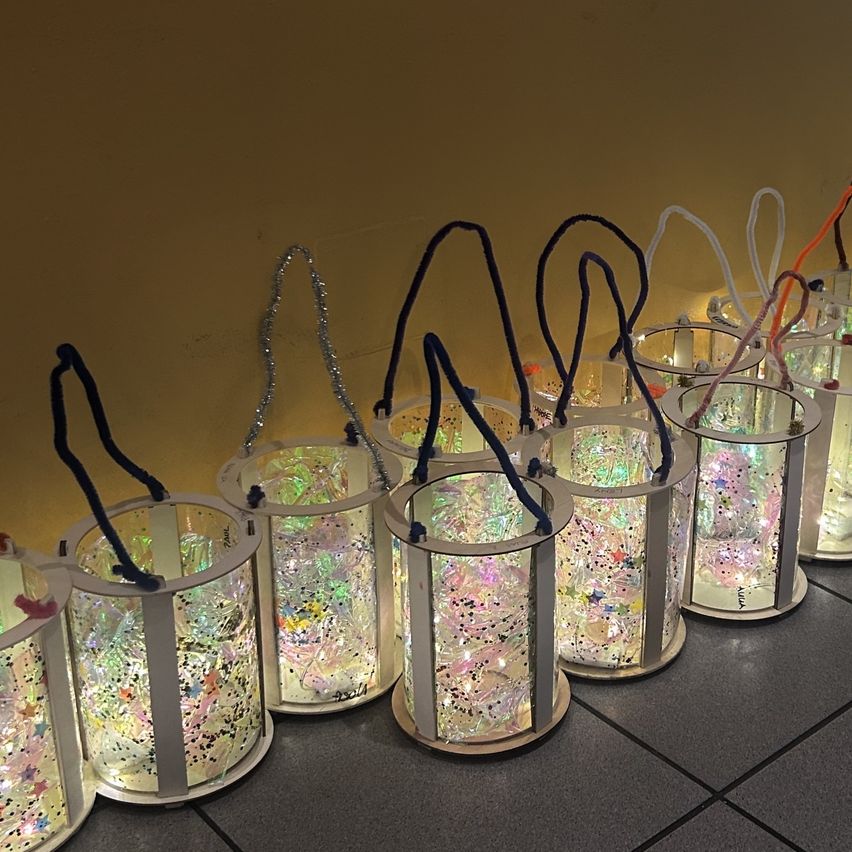 Eine Reihe farbiger Glaslaternen mit Glitzer innen, jede mit einem Griff, aufgereiht auf einem gefliesten Boden an einer gelben Wand.