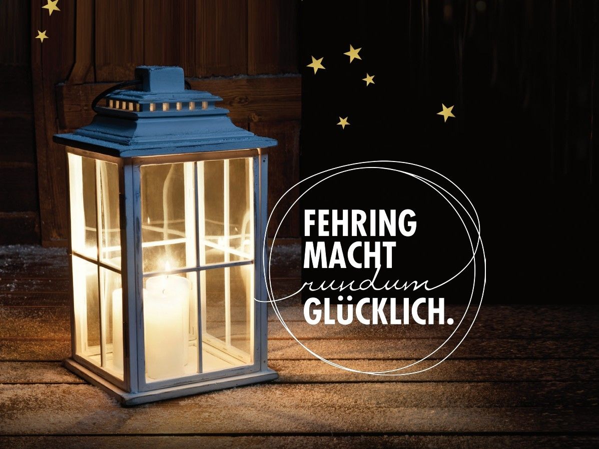 Eine Laterne mit einer brennenden Kerze darin steht auf einer Holzoberfläche. Sie ist von einem Kreis mit dem Text 'FEHRING MACHT RUNDUM GLÜCKLICH' umgeben. Der Hintergrund ist dunkel, und die Laterne ist beleuchtet.