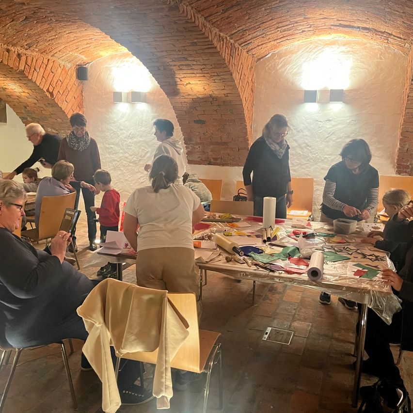 Eine Gruppe von Menschen ist in einem Raum mit Ziegelwänden versammelt, möglicherweise arbeiten sie an einem Bastelprojekt. Einige sitzen, während andere um einen Tisch herum stehen, der mit Papieren, Scheren und einer Rolle Plastik bedeckt ist. Eine Frau sitzt auf der linken Seite und hält ein Tablet.