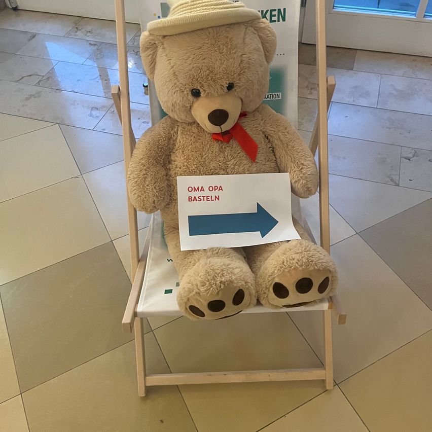 Ein großer Teddybär mit Hut und roter Schleife sitzt auf einem Holzstuhl und hält ein Schild mit einem Pfeil nach rechts.