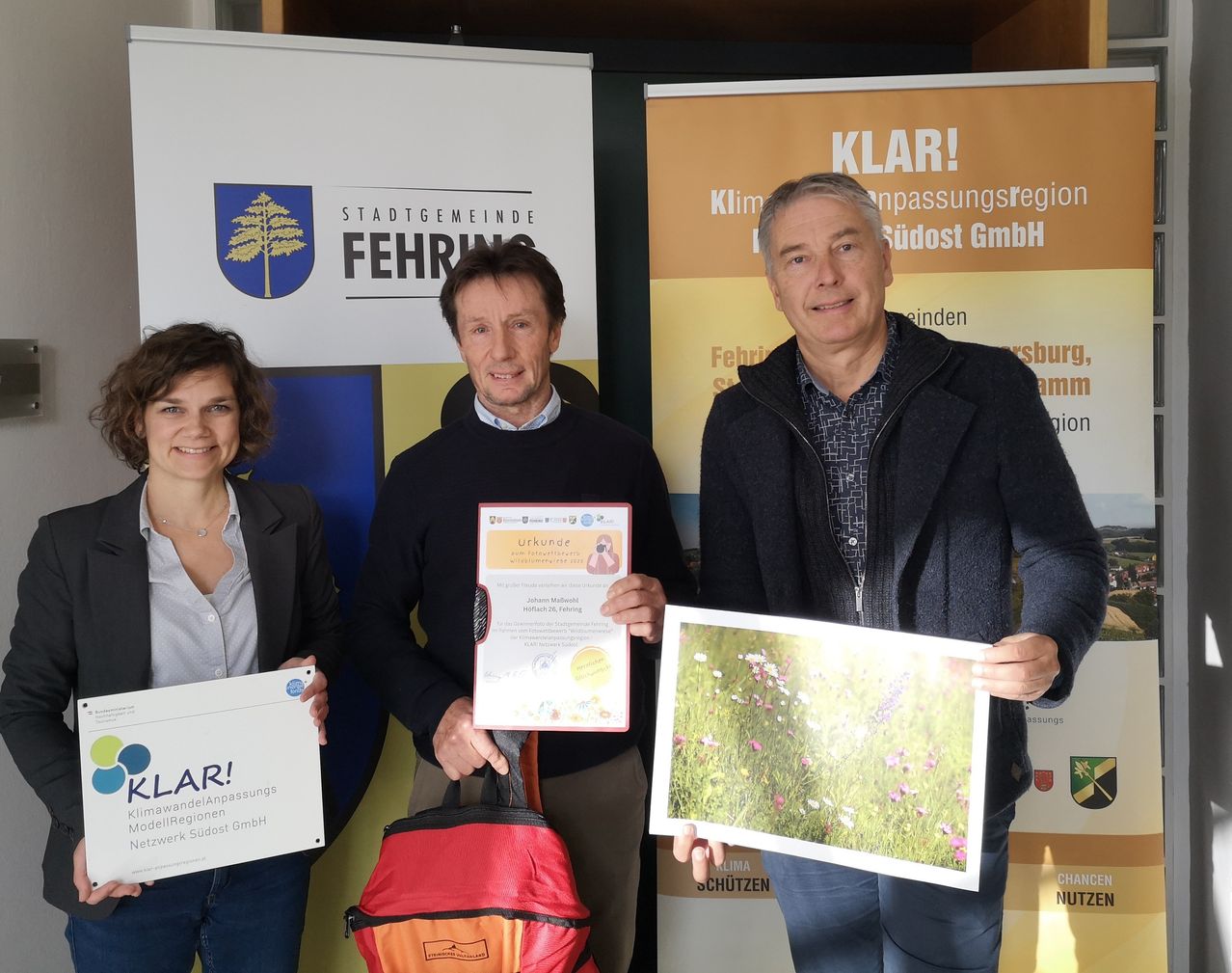 Drei Personen stehen vor einem Banner mit dem Text 'KLAR!' Zwei Männer halten Urkunden und ein Foto, während eine Frau eine Urkunde hält. Sie lächeln und scheinen für ein Foto zu posieren.