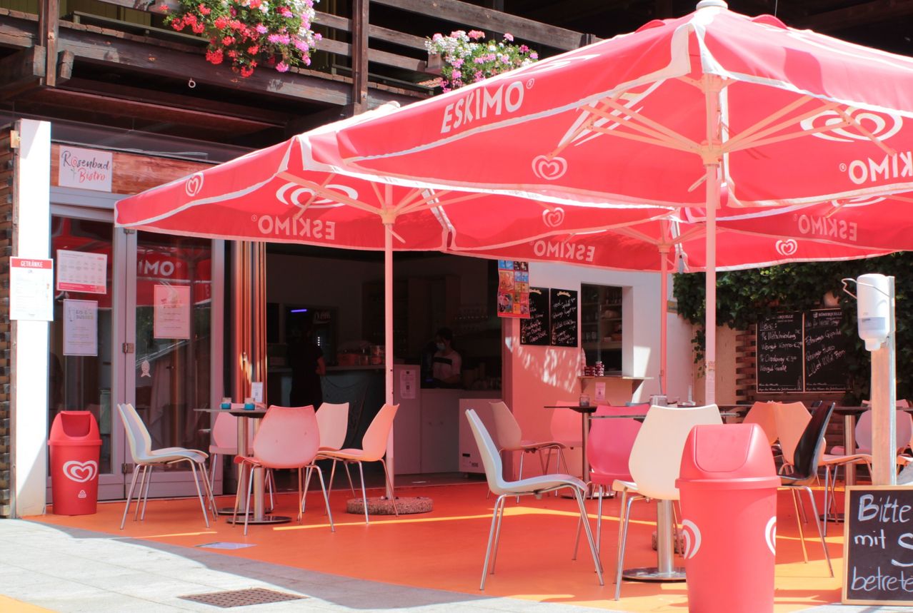 Ein Café mit rosa Sonnenschirmen und Tischen, Stühlen und Mülleimern. Die Sonnenschirme haben das Wort Eskimo darauf. Hinter dem Café steht ein Gebäude mit rosa Wänden.