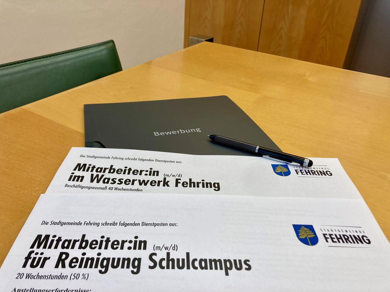 Eine Stellenausschreibung für Mitarbeiter im Wasserwerk und Reinigungsschulcampus liegt auf einem Schreibtisch, zusammen mit einer Mappe mit der Aufschrift Bewerbung. Ein Stift liegt auf der Stellenausschreibung.