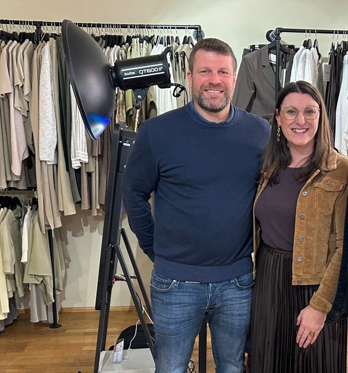 Ein Mann und eine Frau stehen in einem Bekleidungsgeschäft und lächeln in die Kamera. Dahinter befinden sich Kleiderständer und eine große Studioleuchte.