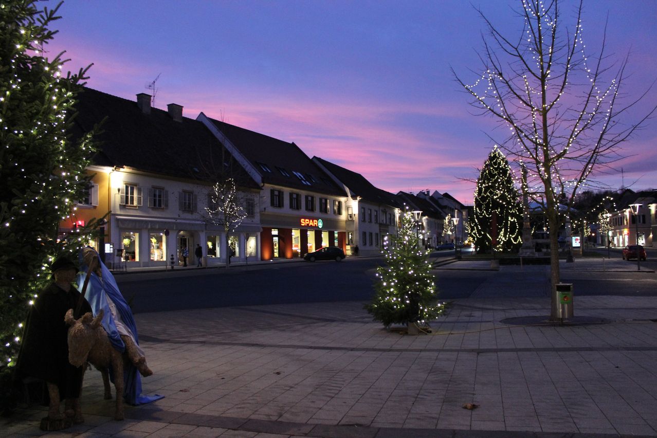 Eine ruhige Straße in der Dämmerung mit Gebäuden, einem beleuchteten Weihnachtsbaum und einem geparkten Auto. Der Himmel hat Töne von Lila und Rosa.