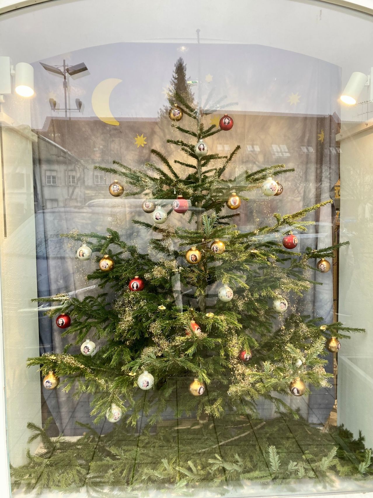 Ein mit roten und goldenen Ornamenten geschmückter Weihnachtsbaum wird in einem Glasfenster ausgestellt. Der Baum ist mit mehreren Ornamenten und einem Stern oben dekoriert. Der Hintergrund zeigt ein Gebäude mit Mond und Sternen.