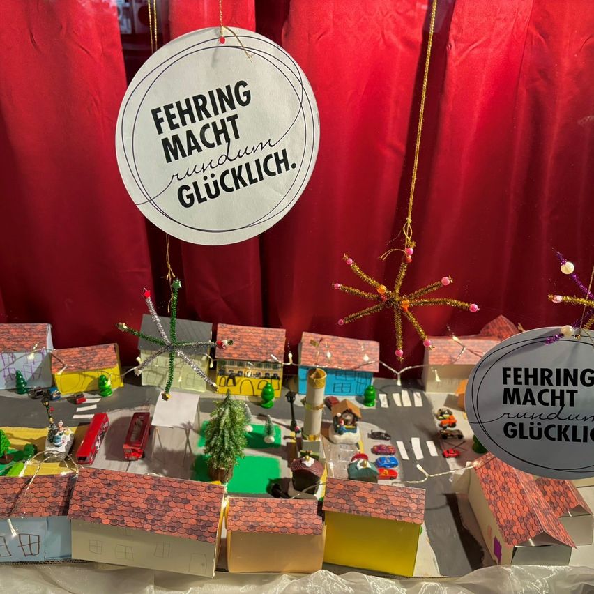 Eine Miniaturmodell einer Kleinstadt mit Häusern und Dekorationen. Zwei Schilder sagen 'Fehriing Macht Rundum Glucklich' auf Deutsch.