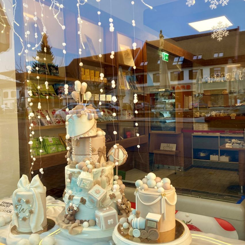 Ein Schaufenster eines Bäckereiladens zeigt zwei mehrstöckige Kuchen, die mit Babyspielzeug, hängenden weißen Schnüren und der Reflexion eines Ladeninneren mit Regalen und Produkten geschmückt sind.