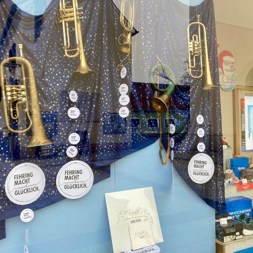 Eine blaue Ausstellung zeigt mehrere Trompeten und Posaunen, die von der Decke hängen. Jedes Instrument ist mit weißen runden Aufklebern versehen, auf denen 'Fehling Macht Glucklich' steht. Darunter befindet sich ein weißes Papier mit Zeichnungen und dem Text 'Soko Dixie'. Rechts sind Schuhe in einer blauen Box ausgestellt.