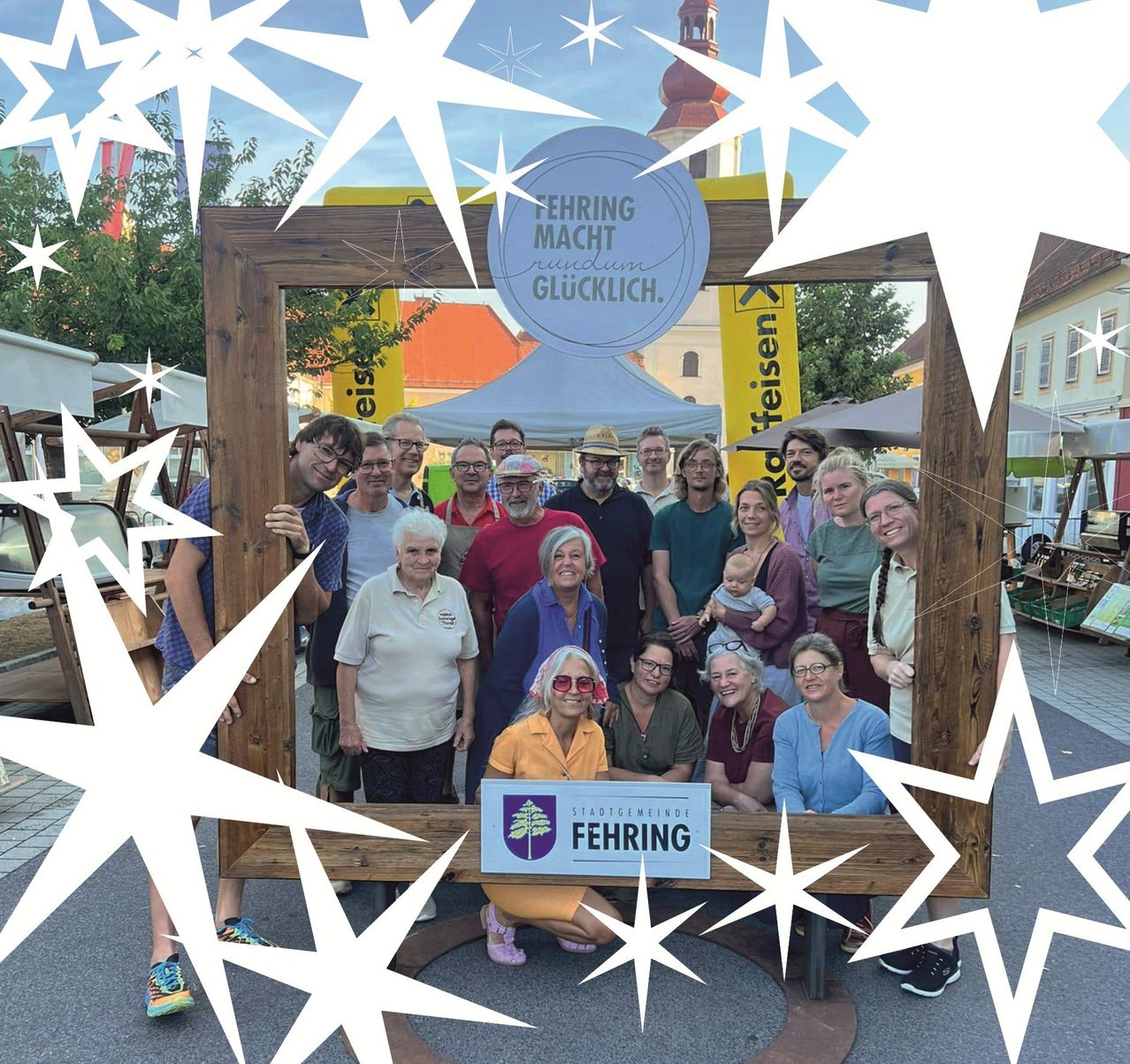 Eine Gruppe von Menschen steht vor einem Holzrahmen mit Sternen und lächelt für ein Foto. Der Rahmen hat deutschen Text und ein Logo. Dahinter stehen Zelte, Bäume und Gebäude.
