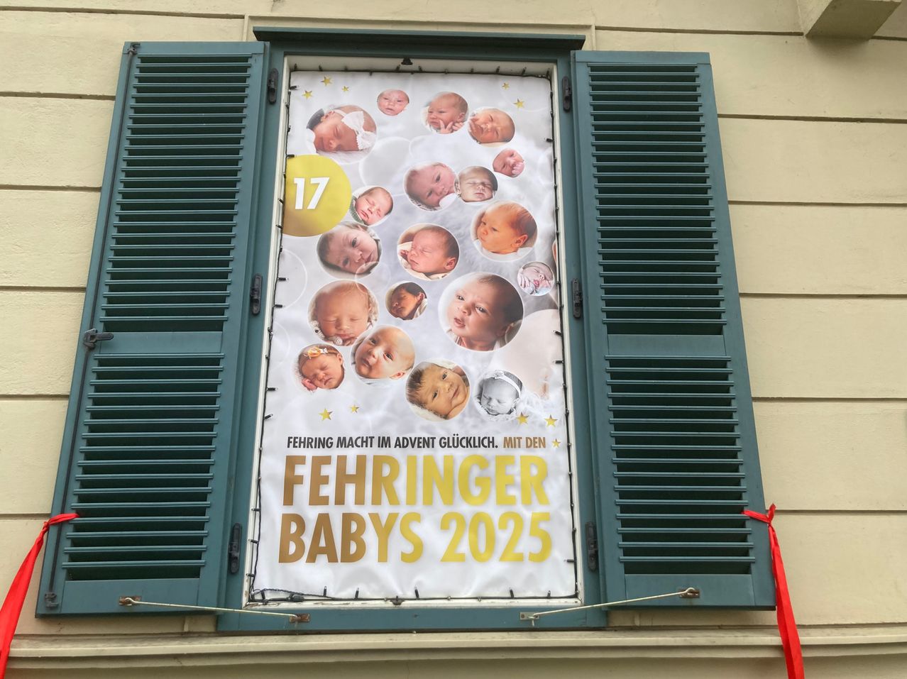 Ein Plakat am Fenster mit 17 Babybildern und dem Text 'Fehringer Babies 2025' in goldenen Buchstaben. Das Plakat ist auf einem beigen Hintergrund.