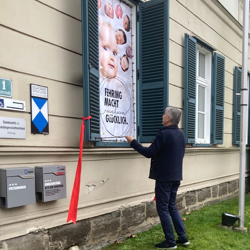 Ein Mann schneidet das Band an einem Gebäude mit einem Plakat mit Babys. Das Plakat sagt 'FEHRING MACHT RUNDUM GLUCKLICH'. Es gibt zwei Briefkästen und ein Schild in der Nähe.