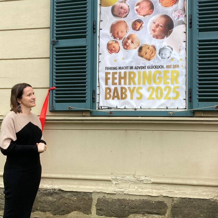 Eine Frau steht neben einem Banner mit Fotos von Säuglingen, betitelt 'Fehringer Babies 2025'. Das Banner ist über einem offenen Fenster mit grünen Fensterläden angebracht.