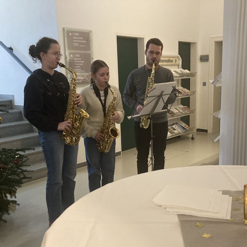 Drei Personen stehen in einem Raum, jede mit einem Saxophon. Vor ihnen steht ein Tisch mit einer weißen Tischdecke und einer Kerze. Hinter ihnen befindet sich eine Treppe mit einem Weihnachtsbaum.