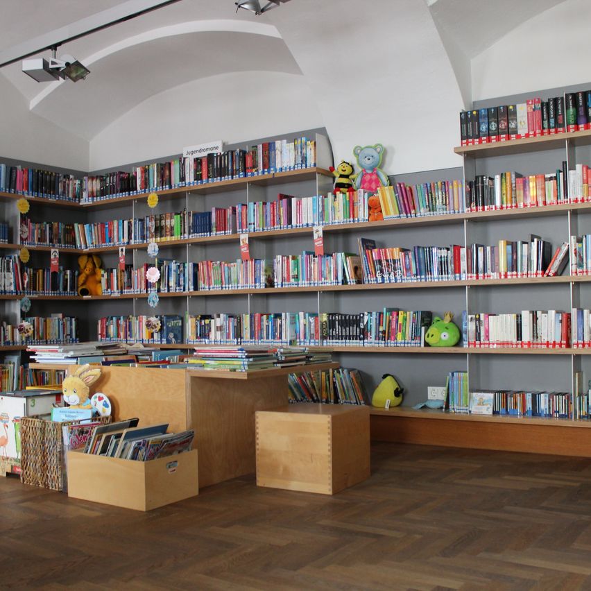 Eine große Bibliothek mit Büchern auf mehreren Regalen. Gefüllte Tiere sind verstreut und ein Schreibtisch mit einem Bücherkorb im Vordergrund.