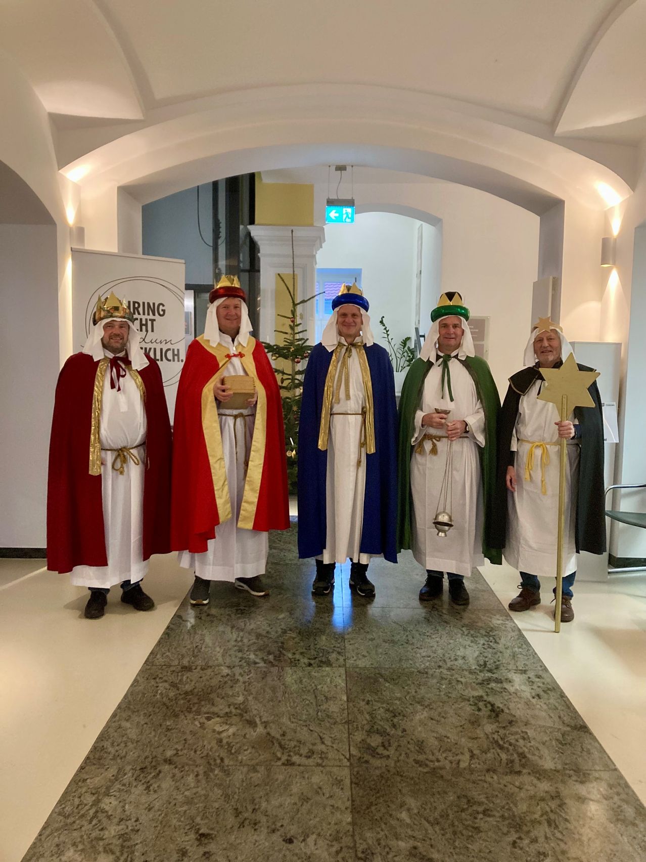 Fünf Männer in mittelalterlichen Kostümen stehen in einem Flur. Einer hält einen Stern und ein anderer eine Schachtel. Sie spielen wahrscheinlich biblische Figuren.