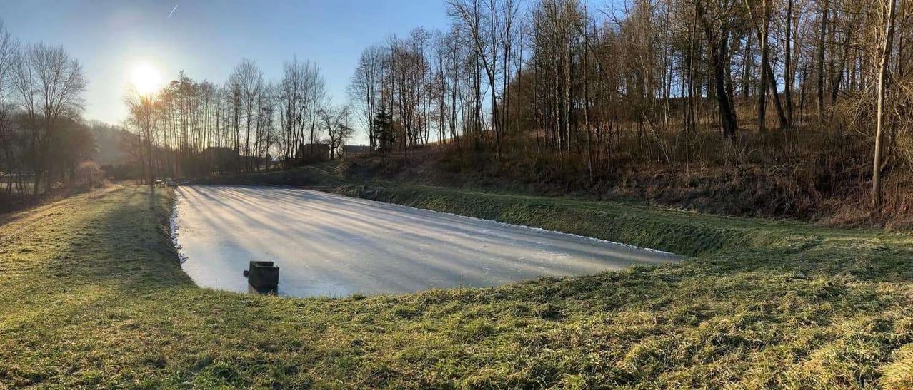 Ein gefrorener Fluss mit einem kleinen Objekt im Wasser, umgeben von Bäumen und einem Hügel. Der Himmel ist klar und blau.