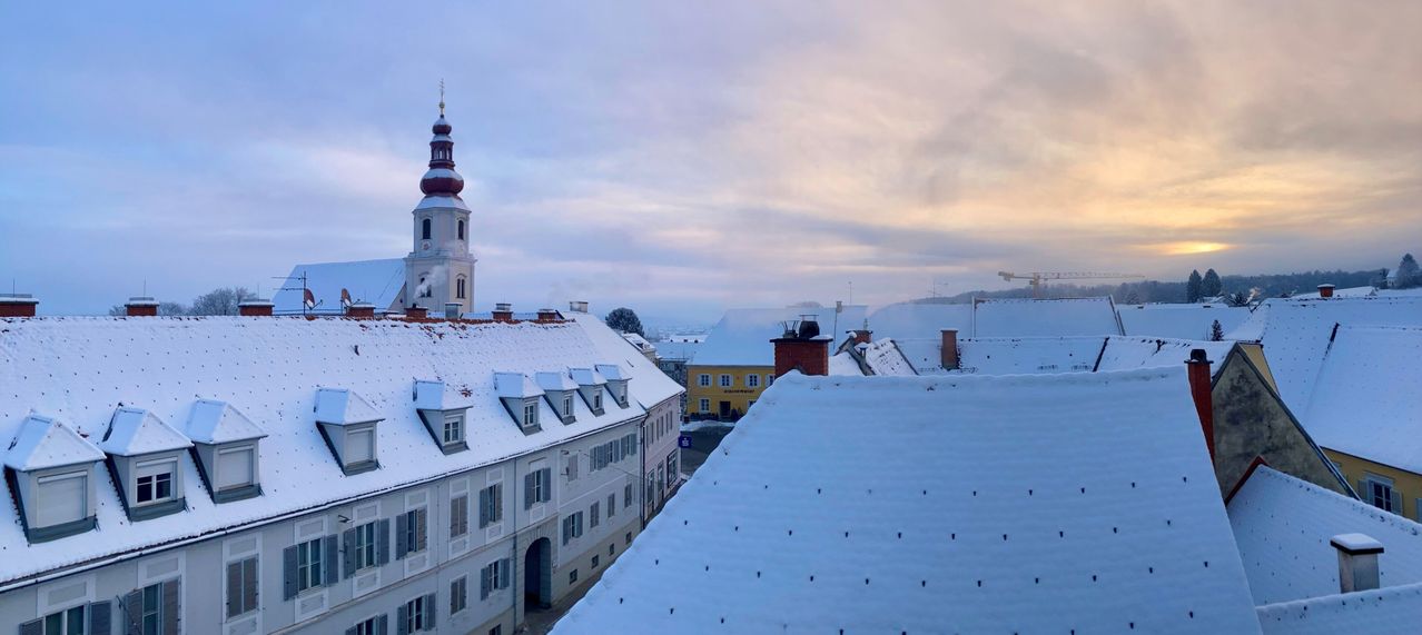 Eine verschneite Stadtlandschaft mit Glockenturm, Schornsteinen und einem gelben Gebäude. Der Himmel ist bewölkt und es liegt Schnee auf den Dächern.