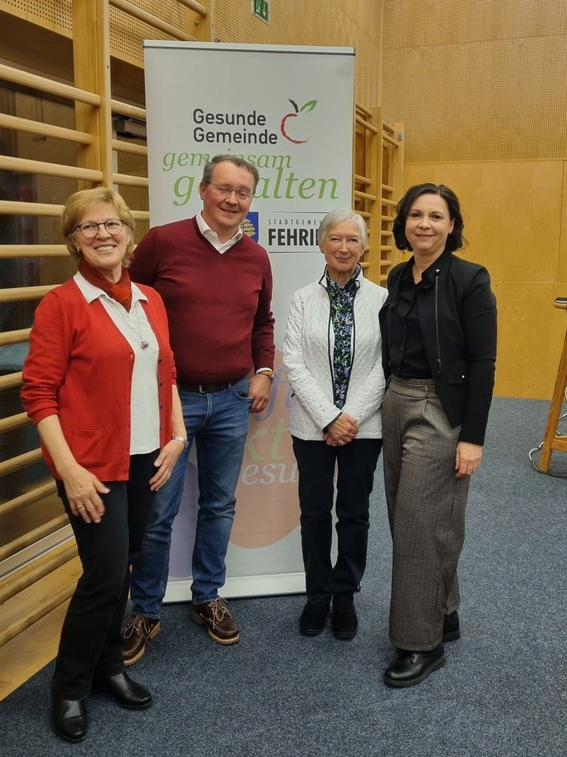 Vier Personen stehen nebeneinander vor einem Banner mit den Worten Gesundheit Gemeinde. Die Frau links trägt eine rote Strickjacke und eine Brille. Der Mann in der Mitte trägt einen bordeauxfarbenen Pullover. Die Frau in der Mitte trägt eine weiße Jacke. Die Frau rechts trägt einen schwarzen Blazer.