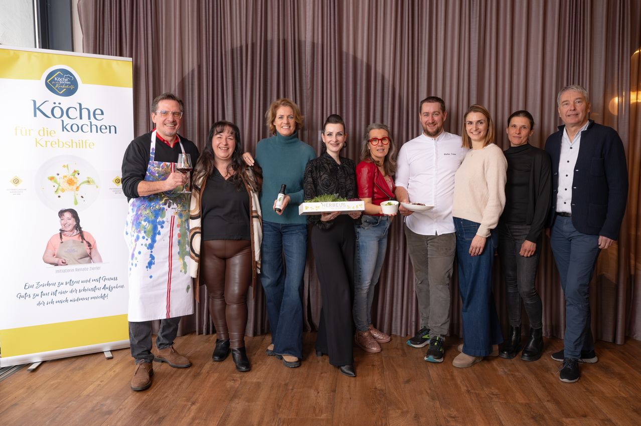 Eine Gruppe von Menschen posiert für ein Foto vor einem Vorhang, lächelt und hält Flaschen und Teller. Die Frau in der Mitte hält eine Box. Hinter ihnen ist ein gelbes Banner.