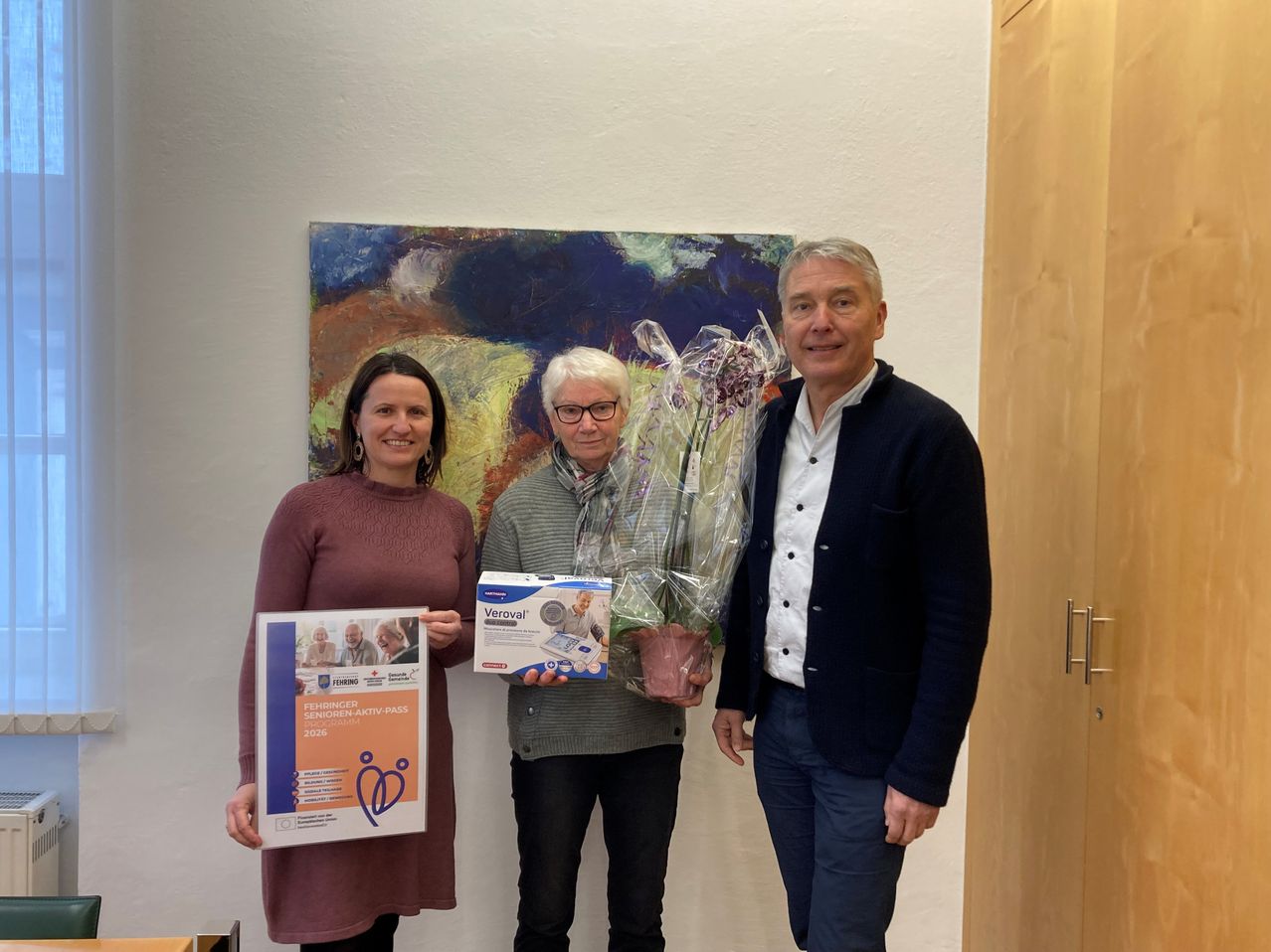 Drei Personen stehen lächelnd vor einem Gemälde. Eine Frau hält ein Plakat, ein Mann einen Blumenstrauß und ein anderer Mann eine Box.