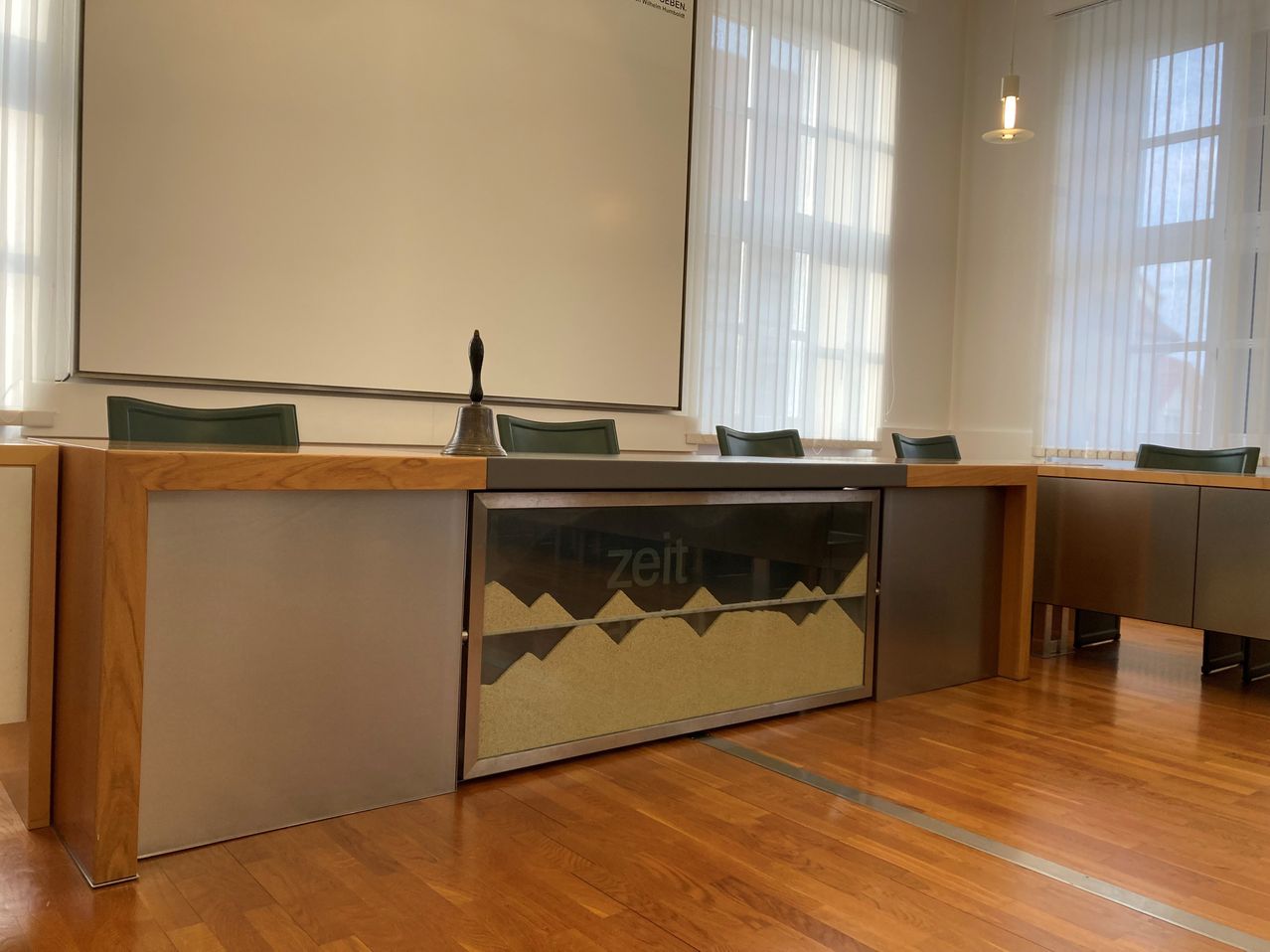 An empty office space with a desk, chairs, and a bell. The desk features a glass partition with the word Zeit. The room has white walls, windows, and a whiteboard.
