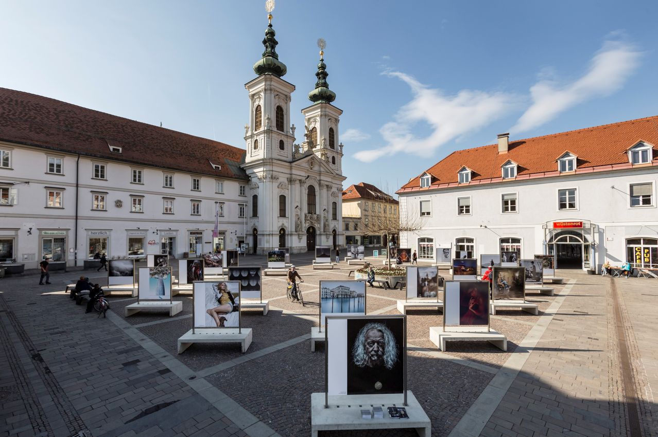 Ein Platz mit gepflastertem Pflaster, der Kunstinstallationen mit verschiedenen Porträts zeigt, vor einer großen weißen Kirche mit zwei Türmen und roten Dächern.