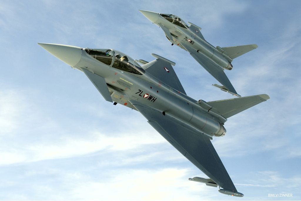 Zwei Kampfjets fliegen am Himmel, einer vor dem anderen. Beide sind grau mit roten Markierungen. Der vordere Jet hat die Buchstaben 'ZLWH' an seiner Seite.