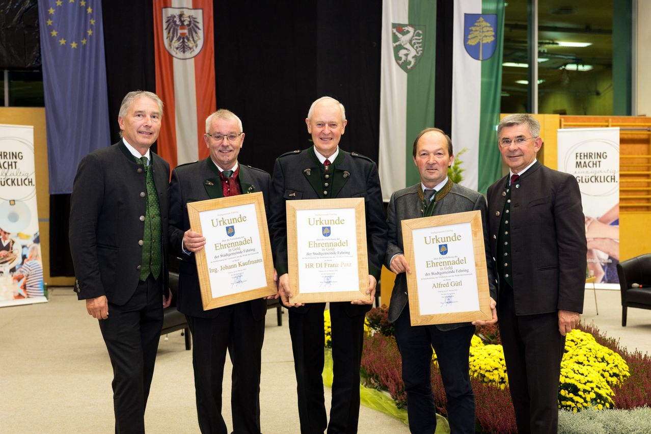 Fünf Männer stehen vor einem Hintergrund mit Flaggen und Blumen und halten Urkunden in Holzrahmen. Der Mann ganz links hält eine Urkunde für Johann Kaufmann. Der zentrale Mann hält eine Urkunde für Franz Patz. Der Mann ganz rechts hält eine Urkunde für Alfred Gill.