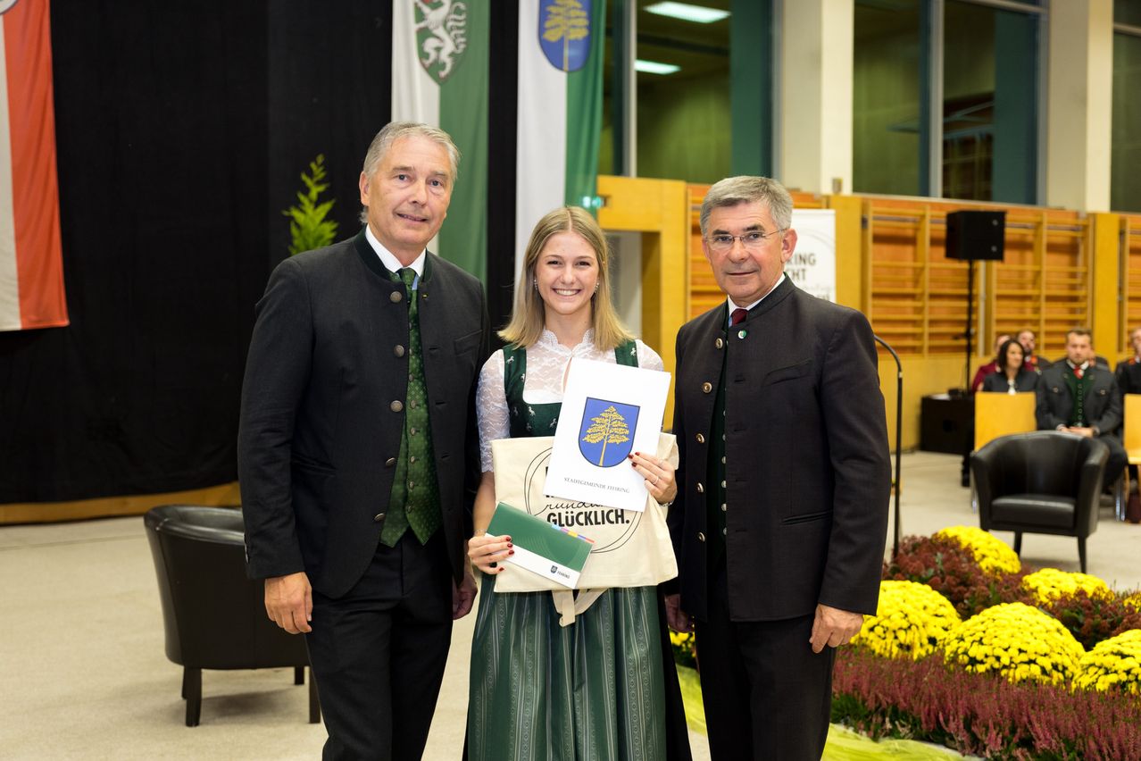 Drei Menschen stehen lächelnd in Innenräumen. Eine Frau in einem grünen Kleid hält ein Zertifikat. Zwei Männer in Anzügen und Krawatten stehen zu ihrer Seite.