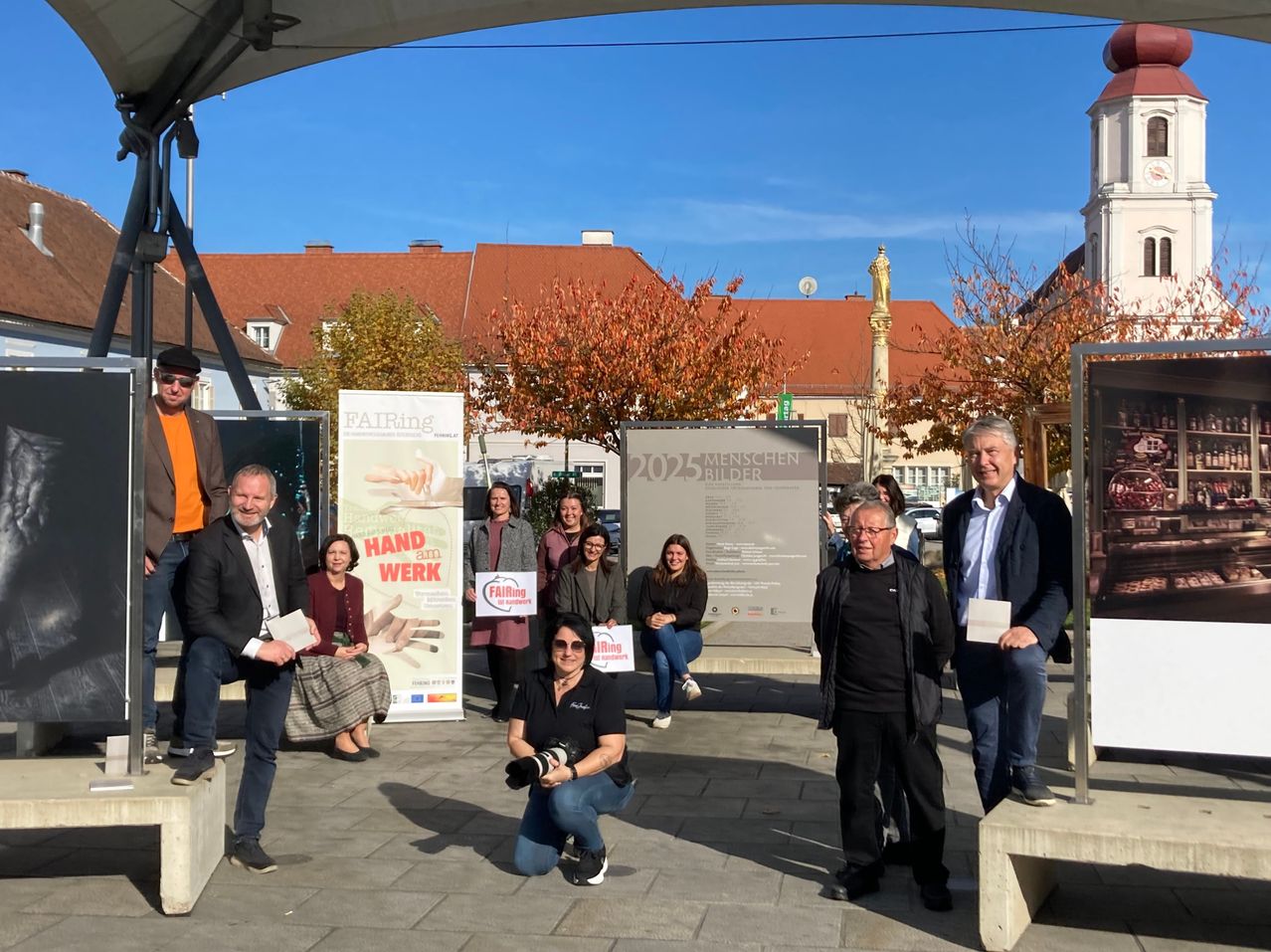 Eine Gruppe von Menschen steht und sitzt auf einer Betonplattform, wahrscheinlich auf einem Stadtplatz. Sie halten Papiere und Schilder. Ein Schild mit dem Text '2025 Menschen' steht hinter ihnen. Hinter dem Schild befinden sich Bäume, ein Turm und Gebäude.