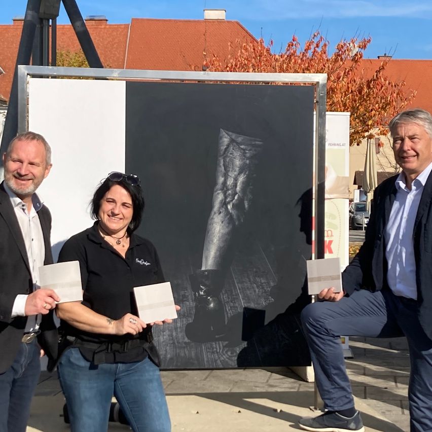 Drei Personen stehen vor einem großen schwarz-weißen Gemälde. Die Frau und der Mann auf der linken Seite halten Papiere, und der Mann auf der rechten Seite lächelt. Dahinter steht ein Gebäude mit roten Dächern.