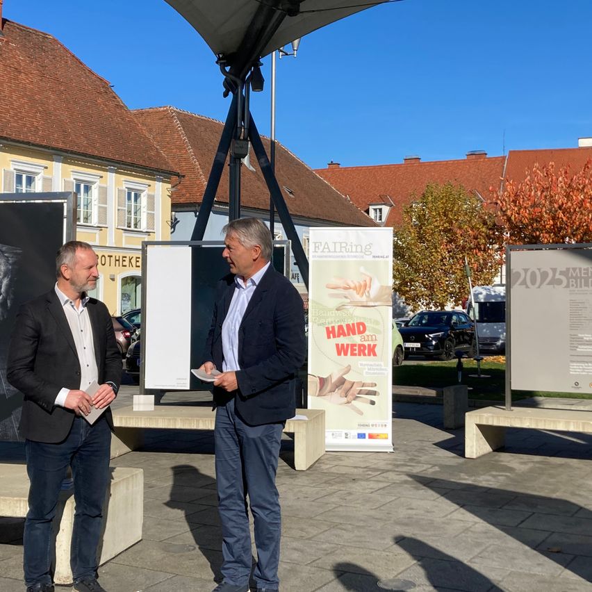 Zwei Männer in Anzügen stehen unter einem Pavillon, einer hält ein Papier, der andere liest. Sie stehen vor einem großen Plakat, das 'Hand am Werk' besagt. Im Hintergrund sind Gebäude und Autos zu sehen.