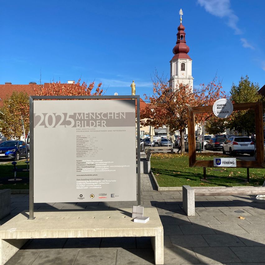 Ein graues Schild zeigt draußen den Titel 2025 Menschen Bilder, mit einem Kirchturm im Hintergrund und mehreren Autos, die am Straßenrand geparkt sind.