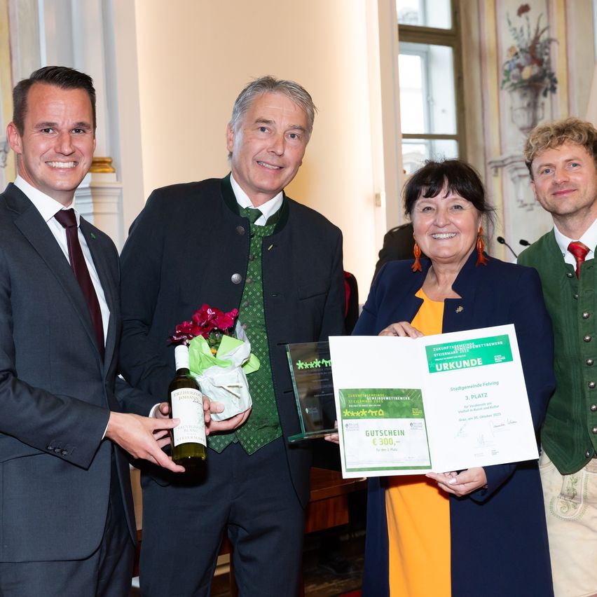 Vier Personen posieren für ein Foto mit Auszeichnungen und Urkunden, einer von ihnen hält eine Weinflasche. Sie stehen in einem Raum mit klassischer Inneneinrichtung.