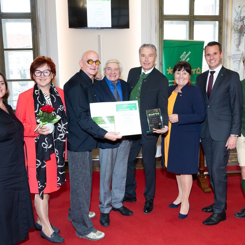 Eine Gruppe von Menschen steht auf einem roten Teppich und hält eine Urkunde. Sie tragen formelle Kleidung. Eine Person hält einen Blumenstrauß. Ein Banner mit Text steht hinter ihnen.