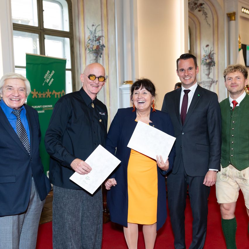 Fünf Personen stehen auf einem roten Teppich, halten Papiere in der Hand und lächeln, mit einem grünen Banner und weißen Säulen im Hintergrund.