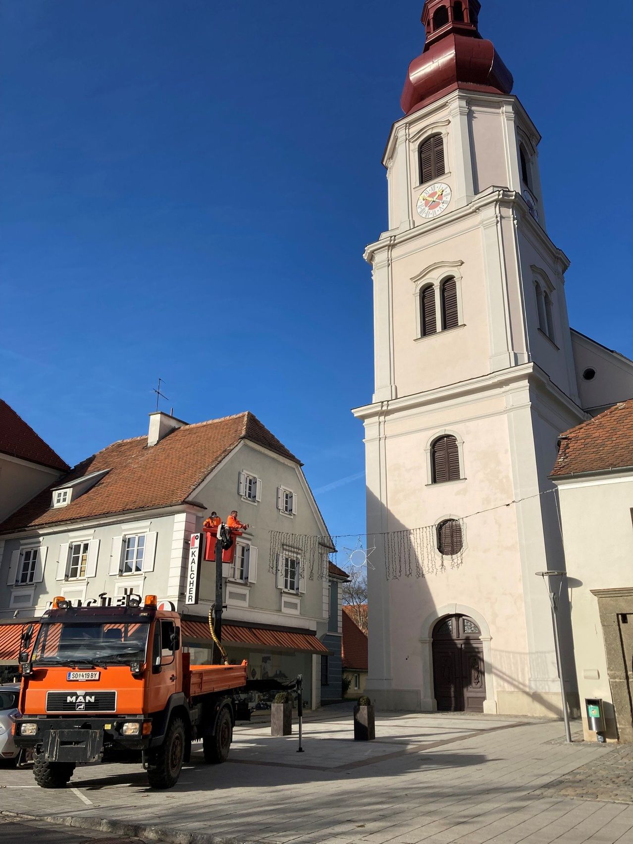 Ein hoher weißer Glockenturm mit einer Uhr auf der Seite steht in der Mitte der Straße. Ein Lastwagen ist vor dem Turm geparkt. Ein Gebäude mit einem braunen Dach befindet sich neben dem Turm. Zwei Personen arbeiten an der Seite des Gebäudes.