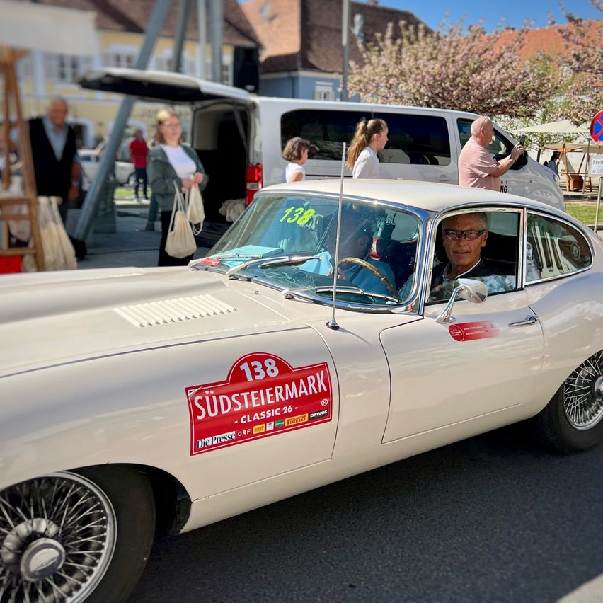 Ein weißer Oldtimer mit einem roten Aufkleber, der 'Südsteiermark' liest, fährt eine Straße hinunter, während Menschen gehen und ein Lieferwagen geparkt ist.