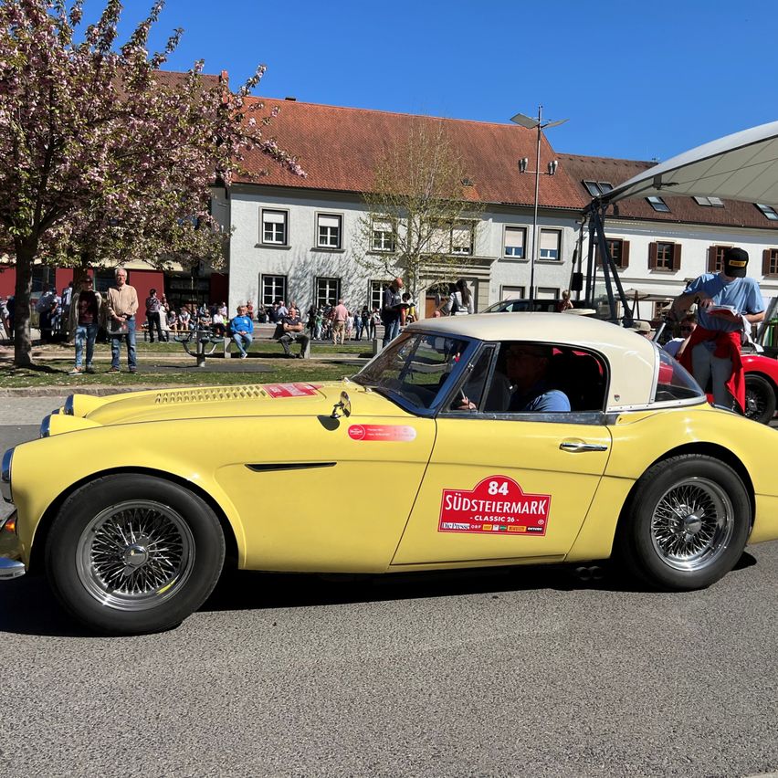 Ein gelber Oldtimer-Sportwagen fährt auf einer Straße, an der eine Gruppe von Menschen steht, in der Nähe eines Gebäudes mit braunem Dach und weißen Wänden.