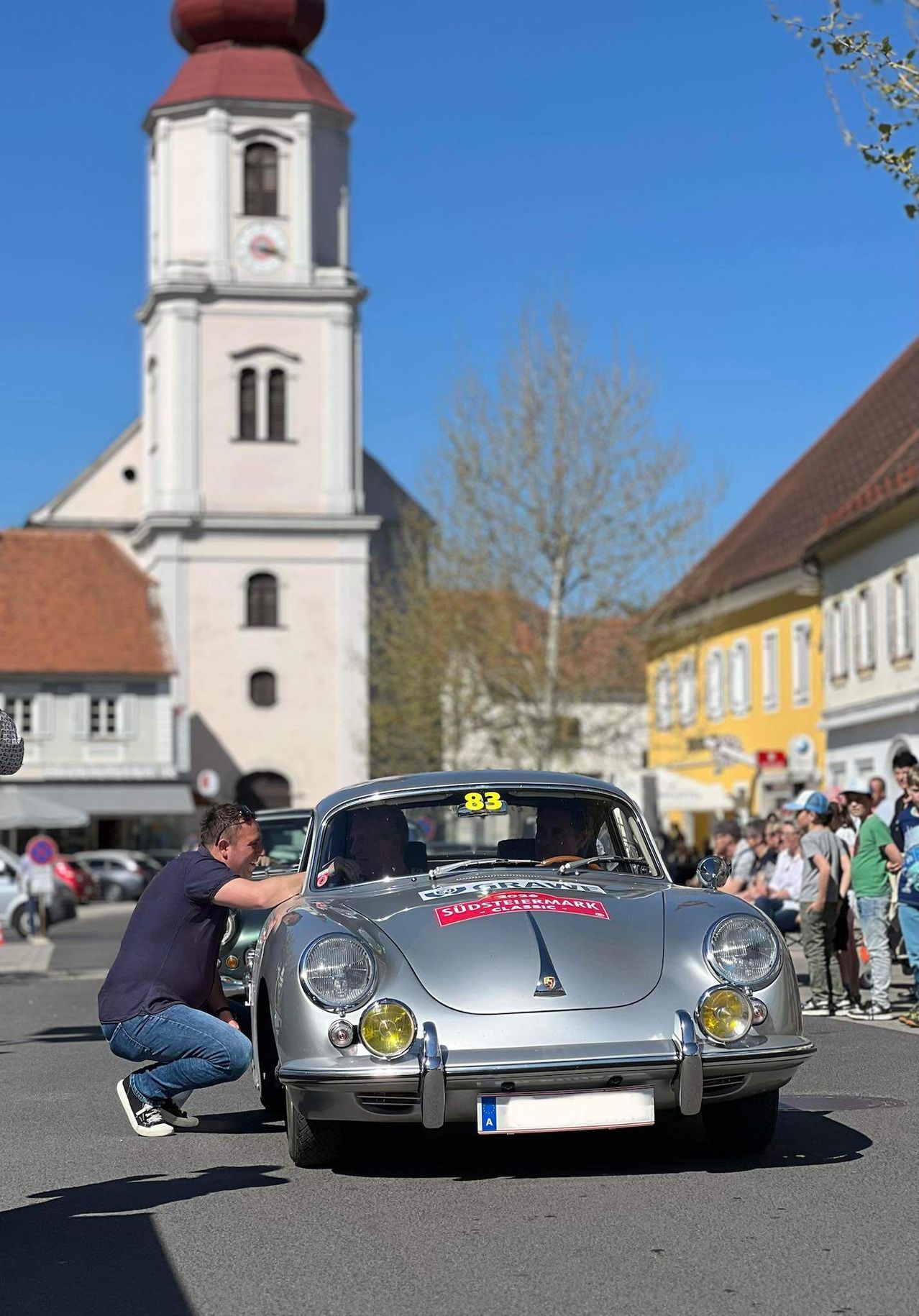 Ein silberner Sportwagen mit der Nummer 83 fährt die Straße entlang, während ein Mann neben ihm kniet. Eine Menschenmenge beobachtet. Im Hintergrund befinden sich Gebäude und ein Baum.