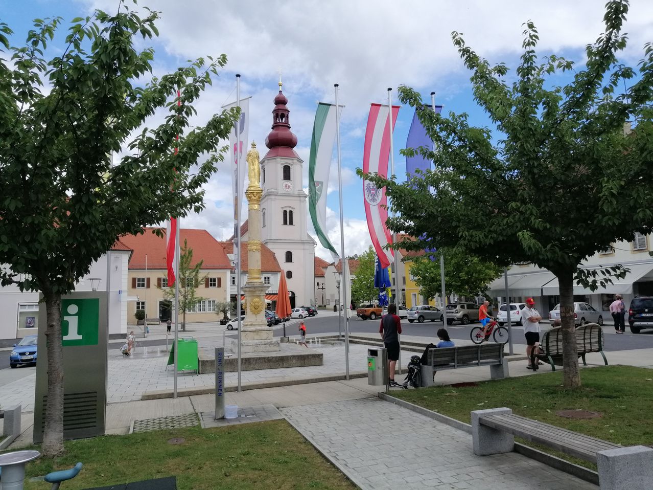 Ein Stadtplatz mit einem Denkmal und Flaggen. Menschen gehen, sitzen auf einer Bank und fahren mit Fahrrädern. Bäume und Gebäude sind in der Umgebung.