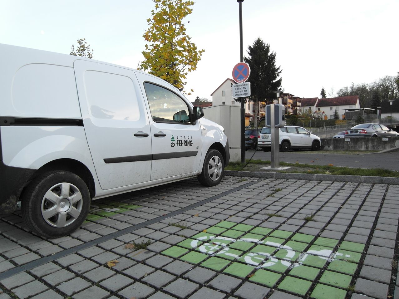 Ein weißer Lieferwagen ist an einer Ladestation auf einem gepflasterten Parkplatz geparkt. Der Lieferwagen trägt ein Schild mit der Aufschrift 'Stadt Fehing.' Im Hintergrund befinden sich Gebäude und Bäume.