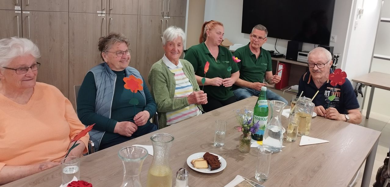 Drei Menschen sitzen an einem Tisch mit einem Teller Essen, Blumen und Getränken. Eine ältere Frau mit weißen Haaren lächelt und hält eine Blume. Eine Frau in Grün hält eine Blume und ein Mann sitzt neben ihr.