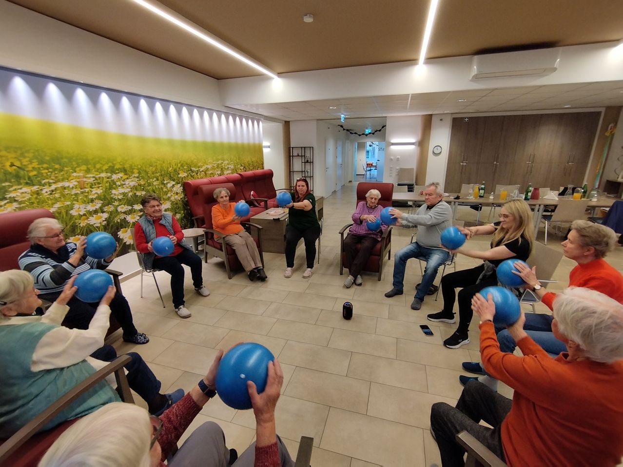A group of older adults is participating in an indoor exercise session. They are seated in chairs, holding blue exercise balls. The room has a mural and is brightly lit.
