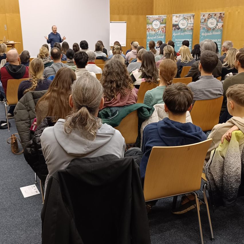 A lecture is being given in a large room, with a man speaking and a white board behind him. The audience, consisting of adults and young people, is seated in chairs.