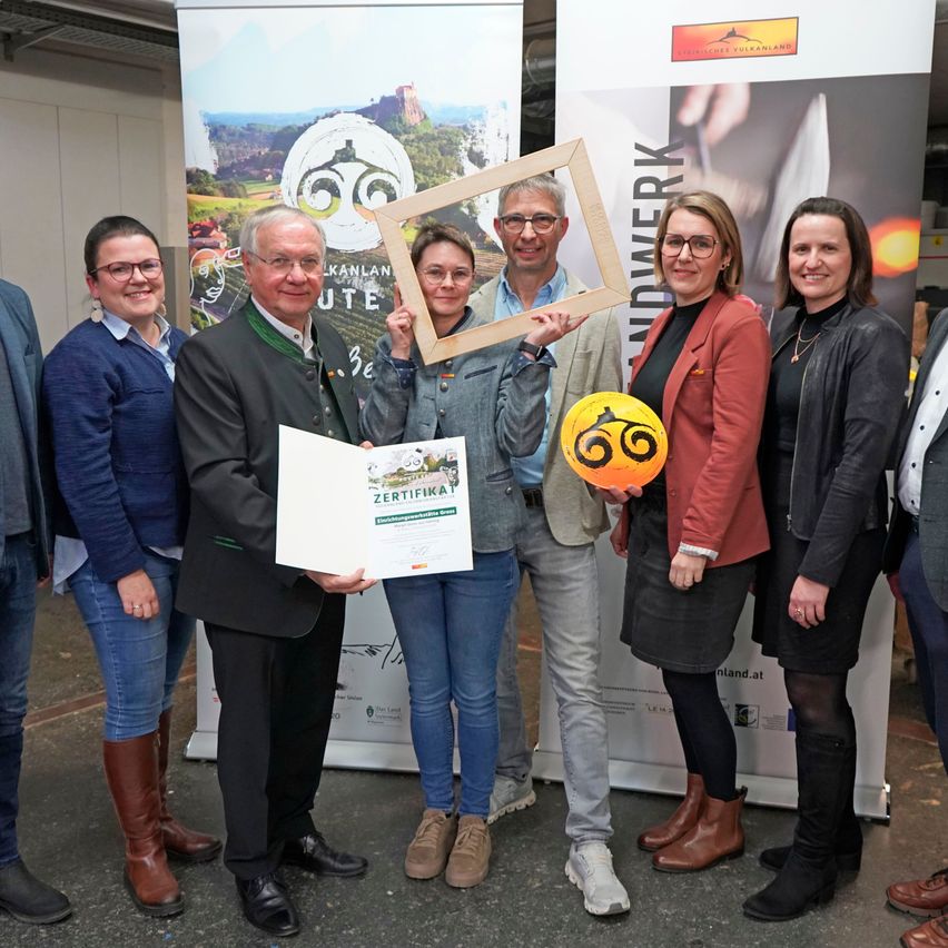 A group of people stand smiling in front of a banner. A man holds a certificate and a frame, while a woman holds a ball. They are all dressed formally.