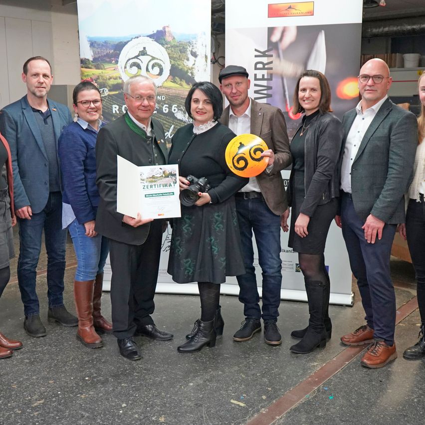 A group of people stand in front of a banner, some holding a certificate and a ball. They are dressed formally and smiling, possibly for a photo.