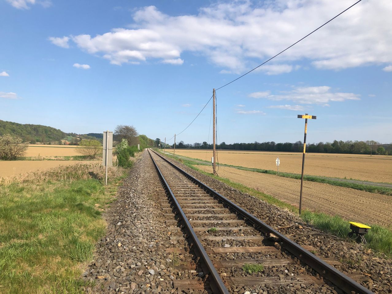 A train track runs through a field with a clear blue sky and some clouds. The track is surrounded by gravel and has poles along its sides.