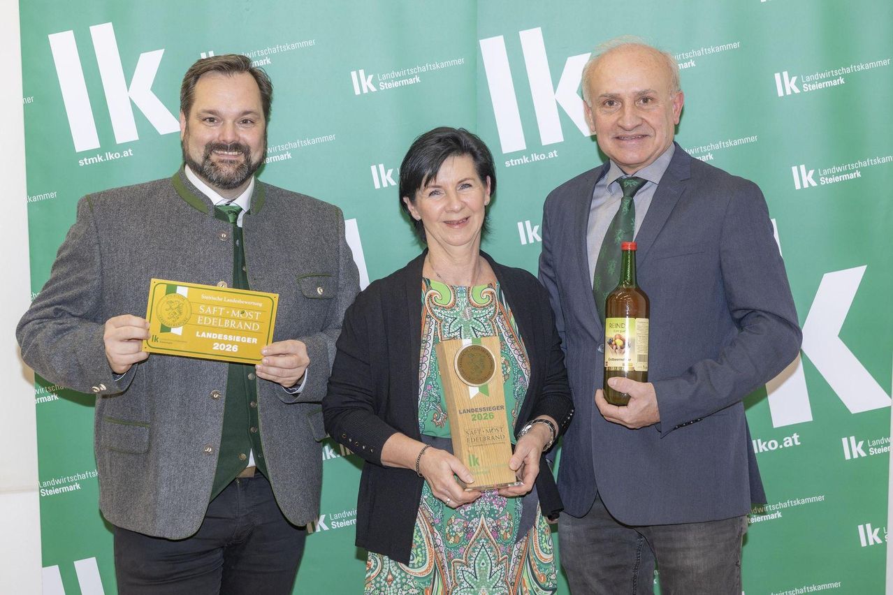 Three people stand in front of a green banner with awards, smiling. A man holds a yellow certificate, a woman a wooden award, and another man a glass bottle.